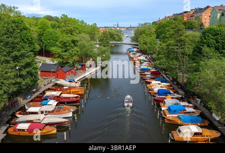 Stockholm, Schweden - 5. Juni 2025: Malerischer Blick auf einen Kanal, der von farbenfrohen Booten gesäumt ist. Üppig grüne Bäume säumen den Wasserweg, und eine Brücke überspannt sich Stockfoto