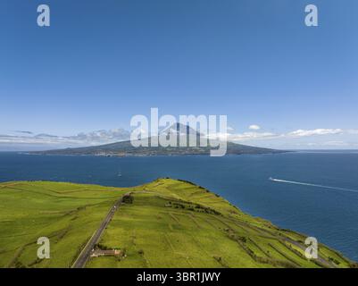 Die majestätische Insel Pico erhebt sich aus dem tiefblauen Atlantik, im Kontrast zur lebhaften grünen Küste im Vordergrund, Horta, Azoren, Portugal. Stockfoto