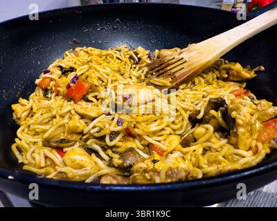 Gebratene Nudeln mit Huhn und Gemüse, die in einem Wok oder einer Pfanne mit Holzspachtel gekocht werden und ein appetitliches Gericht der asiatischen Küche zeigen Stockfoto