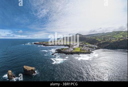 Aus der Vogelperspektive auf die zerklüftete Küste, die ein malerisches Dorf am Fuße der sanften grünen Hügel umschließt, Mosteiros, Azoren, Portugal. Stockfoto