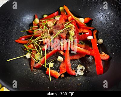 Nahaufnahme von geschnittenen roten Paprika, Zitronengras, gehackten Frühlingszwiebeln in einem schwarzen Wok, gewürzt mit Gewürzen in einer Kochzubereitung Stockfoto