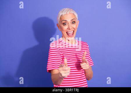 Energiegeladene ältere Frau in einem gestreiften Hemd, die vor auffälligem lila Hintergrund warm lächelt und fröhlich gespendet Stockfoto
