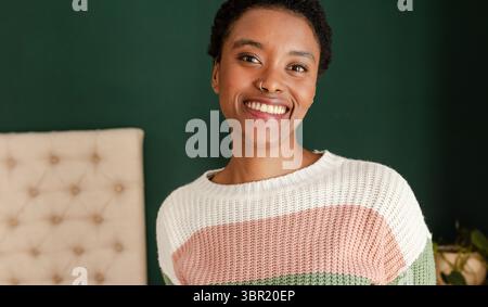 Afroamerikanerin lächelt in die Kamera im Schlafzimmer neben dem Kopfteil mit Topfpflanzen, Kopierraum. Porträt, Wellness, gemütlich, Wohnkultur, natürliches Licht Stockfoto