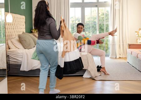 Verschiedene weibliche Freunde halten ein Kleid in Blockfarben und eine passende Hochabels-Sandale auf dem Bett im Schlafzimmer. Mode, Freundschaft, Lifestyle, Eleganz, Dekor, kol Stockfoto
