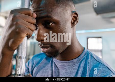 Afroamerikanischer Mann in blauem T-Shirt, der den Metallgriff der Langhantel und den Kopf im Fitnessstudio fasst. Fitness, Kraft, Entschlossenheit, Leichtathletik, Training, E Stockfoto