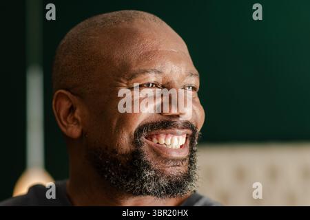 Afroamerikanischer Mann lächelt und blickt direkt im Schlafzimmer mit dunkelgrüner Wand, Lampe und Kopfteil. Porträt, Glück, Wellness, Entspannung, Wärme Stockfoto