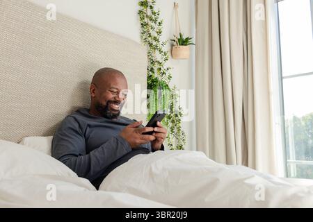 Afroamerikaner lächelt, während er das Smartphone im Bett mit Pflanzgefäßen und Vorhängen hält, Kopierraum. Entspannung, Wellness, modern, gemütlich, digital, natur Stockfoto