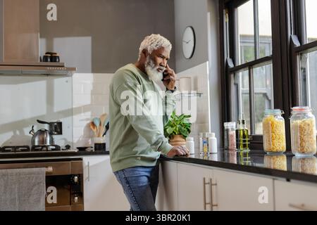 Ranghoher Afroamerikaner, der sich auf die Küchenarbeitsplatte lehnt und mit Pillen telefoniert. Urban, Lifestyle, zeitgenössisch, Gesundheit, Kommunikation Stockfoto