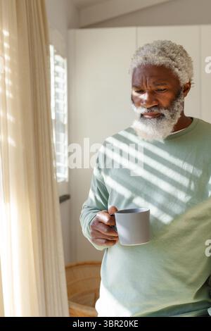 Ranghoher afroamerikanischer Mann, der am verschlossenen Küchenfenster steht und einen grauen Becher im Sonnenlicht hält. Ältere Menschen, Introspektion, Ruhe, Gelassenheit, häuslich Stockfoto