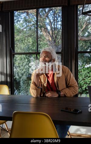 Ranghoher Afroamerikaner sitzt zu Hause am Esstisch unter Bambusjalousien und blickt mit Smartphone. Kontemplation, Introspektion, Ruhe, modern Stockfoto