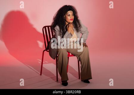 MittelErwachsene afroamerikanische Frau in Bürokleidung, die nachdenklich auf rotem Metallstuhl im Studio sitzt. Kontemplativ, Eleganz, Minimalismus, Raffinesse Stockfoto