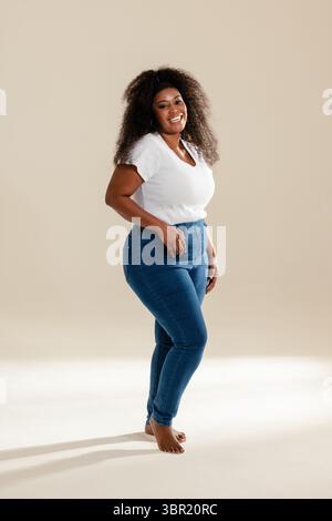 Afroamerikanerin steht barfuß im Studio und trägt ein weißes geripptes T-Shirt und blaue Jeans. Minimalistisch, lässig, Mode, Porträt, Selbstvertrauen, nat Stockfoto