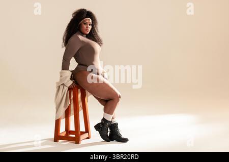 Afroamerikanerin posiert auf dem Hocker mit beigefarbenem Stirnband, cremefarbener Strickjacke im Studio mit Lichtern. Mode, Porträt, minimalistisch, Eleganz, Studio, Stockfoto