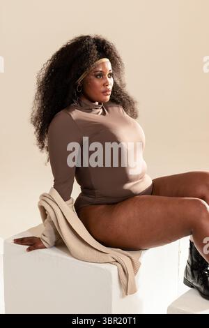 MittelErwachsene afroamerikanische Frau posiert auf weißem Block im Studio mit Stiefeln und Strickjacke. Eleganz, Selbstvertrauen, Mode, Minimalismus, Empowerment, studio, Stockfoto