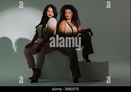 Verschiedene weibliche Kollegen sitzen Rücken an Rücken auf weißen Würfeln im Studio und tragen Hosenträger und Heels. Professionell, modisch, minimalistisch, Porträt, empowe Stockfoto
