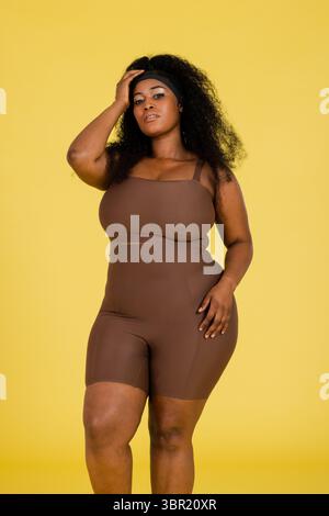 Afroamerikanerin posiert vor gelbem Hintergrund im Studio mit Aktivkleidung und schwarzem Stirnband. Fitness, sportlich, Empowerment, selbstbewusst, modern, Stockfoto