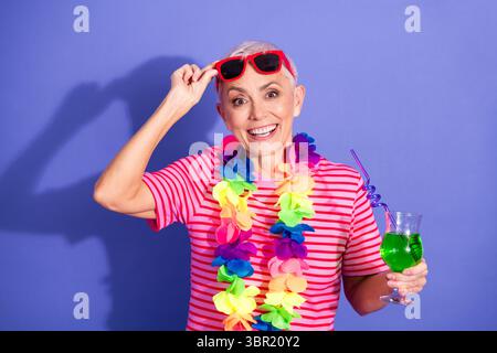 Fröhliche ältere Frau in farbenfroher Sommerkleidung, die ein tropisches Getränk vor einem leuchtend violetten Hintergrund genießt Stockfoto