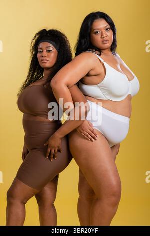 Weibliche Models, die im Studio Rücken an Rücken in brauner und weißer Shapewear mit gelbem Hintergrund stehen. Plus-Größe, Selbstvertrauen, Empowerment, Körperpositivit Stockfoto