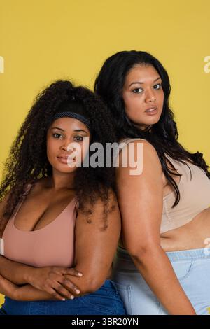 Verschiedene Frauen stehen im Studio zurück an Rücken, tragen rosa und beige Sport-BHs zu Jeans. Mode Models, redaktionell, lebendig, empowering, Casualwea Stockfoto