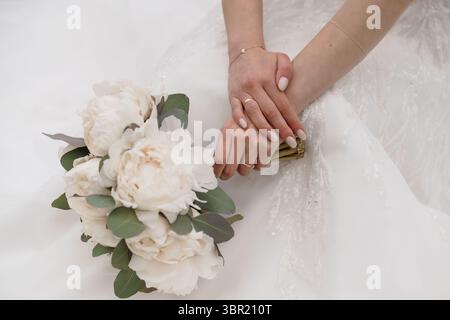Nahaufnahme mit den Händen des frisch vermählten Brautpaares, die einen weißen Pfingstrosenstrauß halten, verziert mit zarten goldenen Trauringen und Armband, romantischer Braut Detai Stockfoto