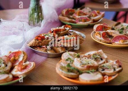 Ein einladender Schuss mit einer Vielzahl von frischen Bruschetta mit Tomaten, Salami, Kräutern und Feta, angeordnet auf Tellern und einem Metalltablett, perfekt für Stockfoto
