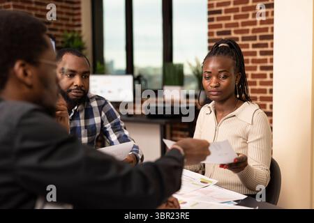 Afroamerikanische Frauen und Männer bewerten Finanzberichte während der Startup-Firmensitzung. Schwarze weibliche Mitarbeiterin, die Finanzdokumente von einem Kollegen in einem stationären Arbeitsbereich erhält. Stockfoto