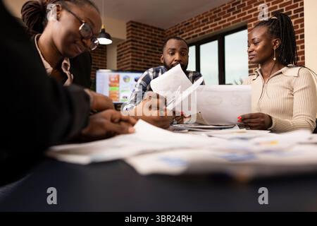 afroamerikanische Kollegen versammelten sich am Tisch und überprüften während der Besprechung zur Bewertung von Startup-Unternehmen mehrere Dokumente. Professionelle schwarze Männer und Frauen analysieren Finanzpläne und Berichte. Stockfoto