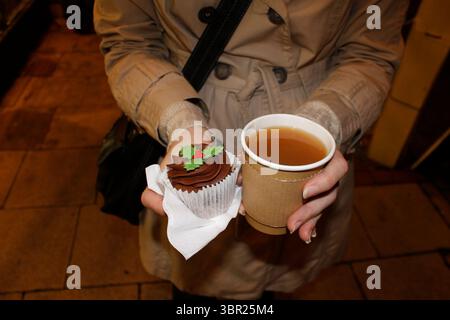 Hände einer Frau, die ein Papercup mit Glühwein und einem Schokoladenmuffin auf einem Weihnachtsmarkt hält. Stockfoto