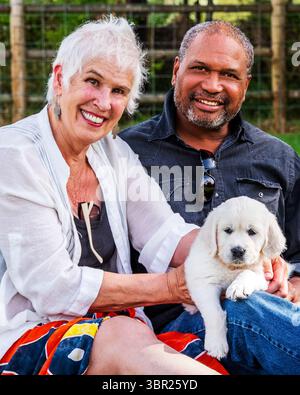 Interrassisches Seniorenpaar mit sechs Wochen altem Platin- oder cremefarbenem Golden Retriever-Welpen. Stockfoto