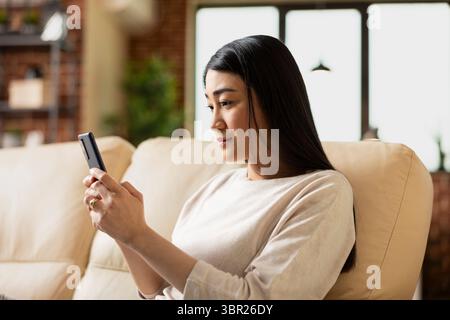 Asiatische Unternehmerin, die zu Hause auf der Couch sitzt, ihr Handy hält und soziale Medien überprüft. Externe Mitarbeiterin, die während der Arbeit von zu Hause aus eine Pause vom Laptop macht. Stockfoto