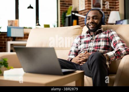 Geschäftsmann macht eine Pause von der Arbeit, sitzt mit Kopfhörern und schaut sich lustige Filme auf Laptop in einem gemütlichen Apartment an. Schwarzer männlicher Freelancer auf dem Sofa lächelt während des Videokonferenzens auf den Bildschirm des Geräts. Stockfoto