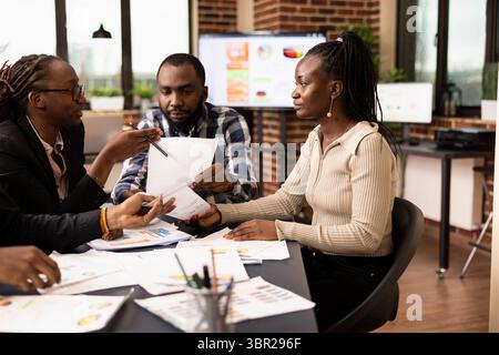 Afroamerikanische Kollegen versammelten sich an einem Konferenztisch und diskutierten und analysierten Finanzberichte. Männliche und weibliche Mitarbeiter mit Dokumenten, Überprüfung der Marketingpläne während der Bewertungsbesprechung. Stockfoto