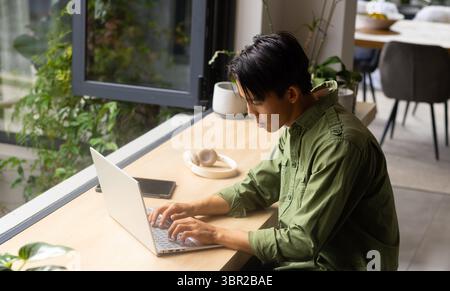 Mann tippt auf Laptop auf hölzerner Arbeitsplatte neben geöffnetem Fenster mit Smartphone-Kopfhörern und Pflanzen. Gemütlich, Produktivität, modern, Arbeitsbereich, Natur, ser Stockfoto