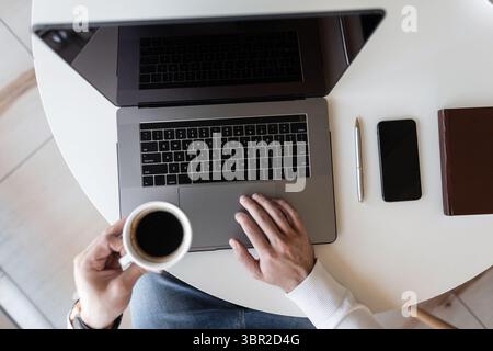 Geschäftsmann, der an Einem Laptop arbeitet und Kaffee trinkt, während er an Einem Tisch in Einem Café sitzt. Der Kerl Arbeitet Aus Der Ferne Am Computer. Draufsicht Auf Dem Desktop. Stockfoto