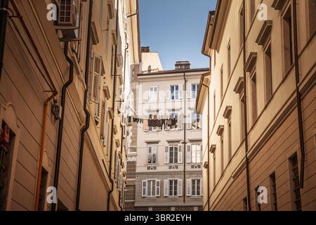 Mit Blick auf eine enge Gasse im historischen Zentrum von Triest in Richtung eines sonnendurchfluteten Innenhof in italien, bilden die Wände eine Kleiderlinie, die zwischen verschlossenen Fenstern angeordnet ist, c Stockfoto