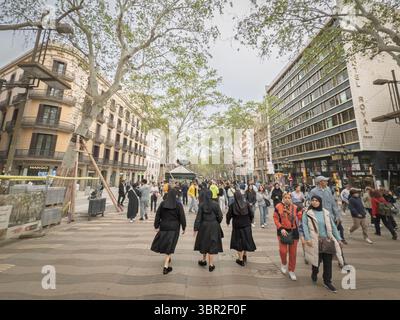 BARCELONA, SPANIEN - 12. APRIL 2025: Drei Nonnen in schwarzen Gewohnheiten gehen die Las Ramblas von Barcelona hinauf und mischen sich mit Touristen, Schildern und Hotels, die die Ramblas umrahmen Stockfoto