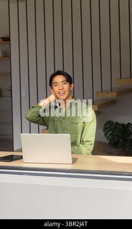 Asiatischer Mann, der den Hals am Schreibtisch im Heimbüro in grünem Hemd mit Laptop-Smartphone-Topfpflanze dehnt Stockfoto