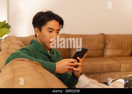Erwachsener asiatischer Mann, der auf Sofa im Wohnzimmer sitzt und Smartphone neben Topfpflanzen hält, Kopierraum Stockfoto