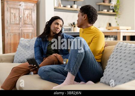 Verschiedene Freundinnen sitzen auf beigefarbenem Sofa mit dekorativen Kissen und halten das Smartphone zu Hause Stockfoto