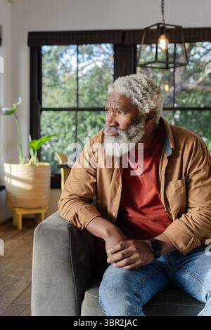 Ranghoher Afroamerikaner sitzt zu Hause in einem grauen Sessel, trägt Smartwatch und blickt ins Fenster Stockfoto