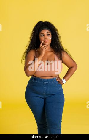 Mittelgroße afroamerikanische Frau zeigt Smartwatch mit Sport-BH und Jeans vor gelbem Hintergrund Stockfoto