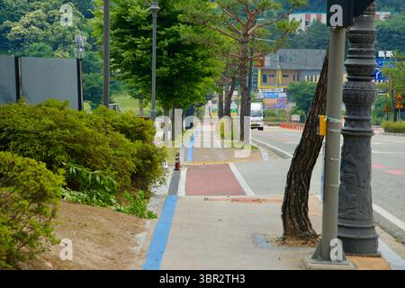 Gongju, Südkorea - 13. Juni 2025: Ein blau lackierter Wanderweg und ein Gehweg führen parallel zur Straße und führen Fußgänger und Radfahrer durch eine schattige Co Stockfoto