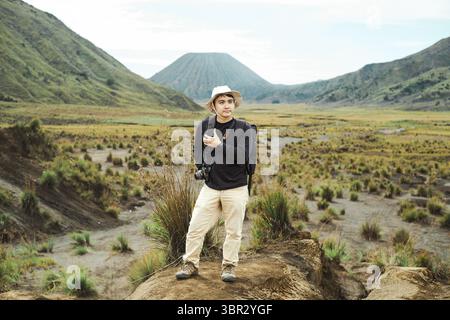 Ein junger südostasiatischer Mann mit Hut und Rucksack steht auf einem Hügel am Mount Bromo, Ost-Java, Indonesien. Kamera in der Hand, bereit zum Erkunden. Perfekt für Werbung Stockfoto