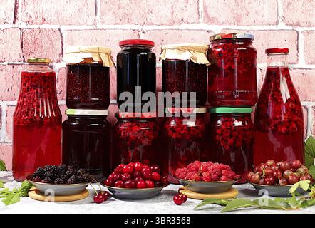 Gläser mit Himbeere, Kirsche, Stachelbeermarmelade und Kompott auf Küchenhintergrund mit Blättern und frischen Beeren, Hauskonservierungskonzept, Banner und Werbung Stockfoto