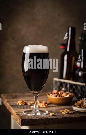 Glas dunkles Bier mit Schaumstoffkopf auf dunklem Holz Hintergrund mit leeren Flaschen und Biersnacks Stockfoto