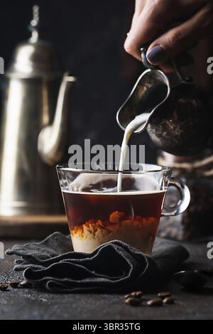 Frau Hand gießen Milch in Glas Tasse Kaffee auf schwarzem Hintergrund Stockfoto