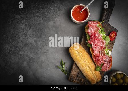 Salchichon extra oder Salami-Sandwich, gegrilltes Getreidebrot, mit Tomaten und Oliven auf dunklem Hintergrund, Draufsicht, Nahaufnahme. Gesundes hausgemachtes Frühstück Stockfoto
