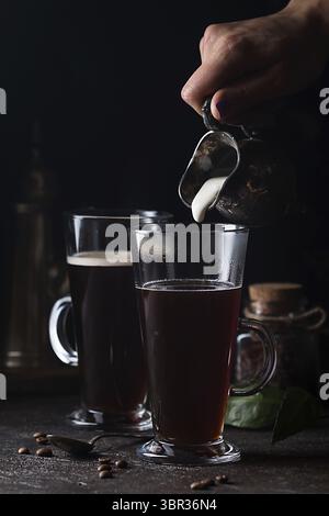 Frau Hand gießen Milch in Glas Tasse Kaffee auf schwarzem Hintergrund Stockfoto