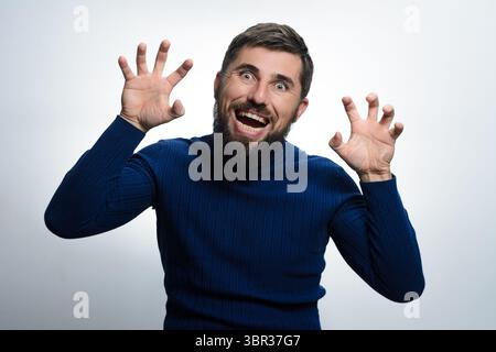 Ein bärtiger Mann in einem blauen Pullover bewegt sich spielerisch mit ausgestreckten Händen zur Kamera. Komischer Ausdruck auf seinem Gesicht gegen einen einfachen Hinterwäldler Stockfoto