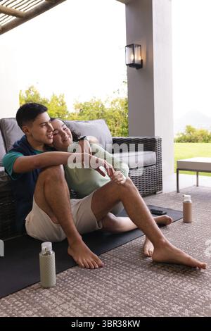 Paare sitzen auf überdachten Terrassen-Yogamatten in Aktivkleidung und lehnen sich an ein Weidensofa mit Wasserflaschen Stockfoto